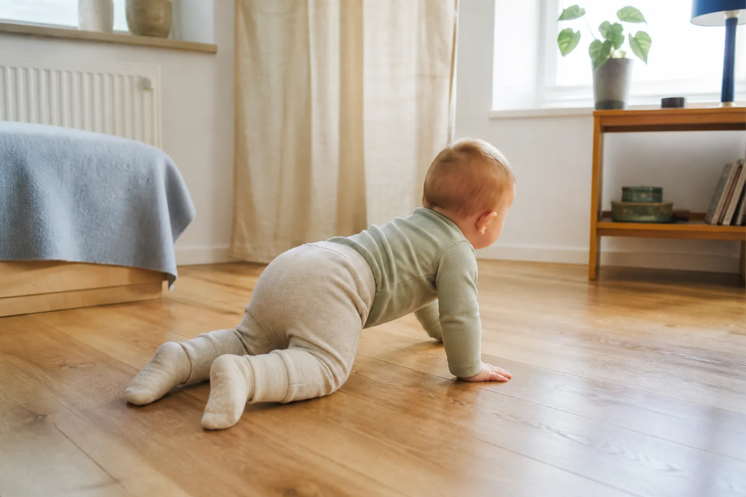 A crawling baby in a pale sage bodysuit, light pants and cream socks on a wooden floor