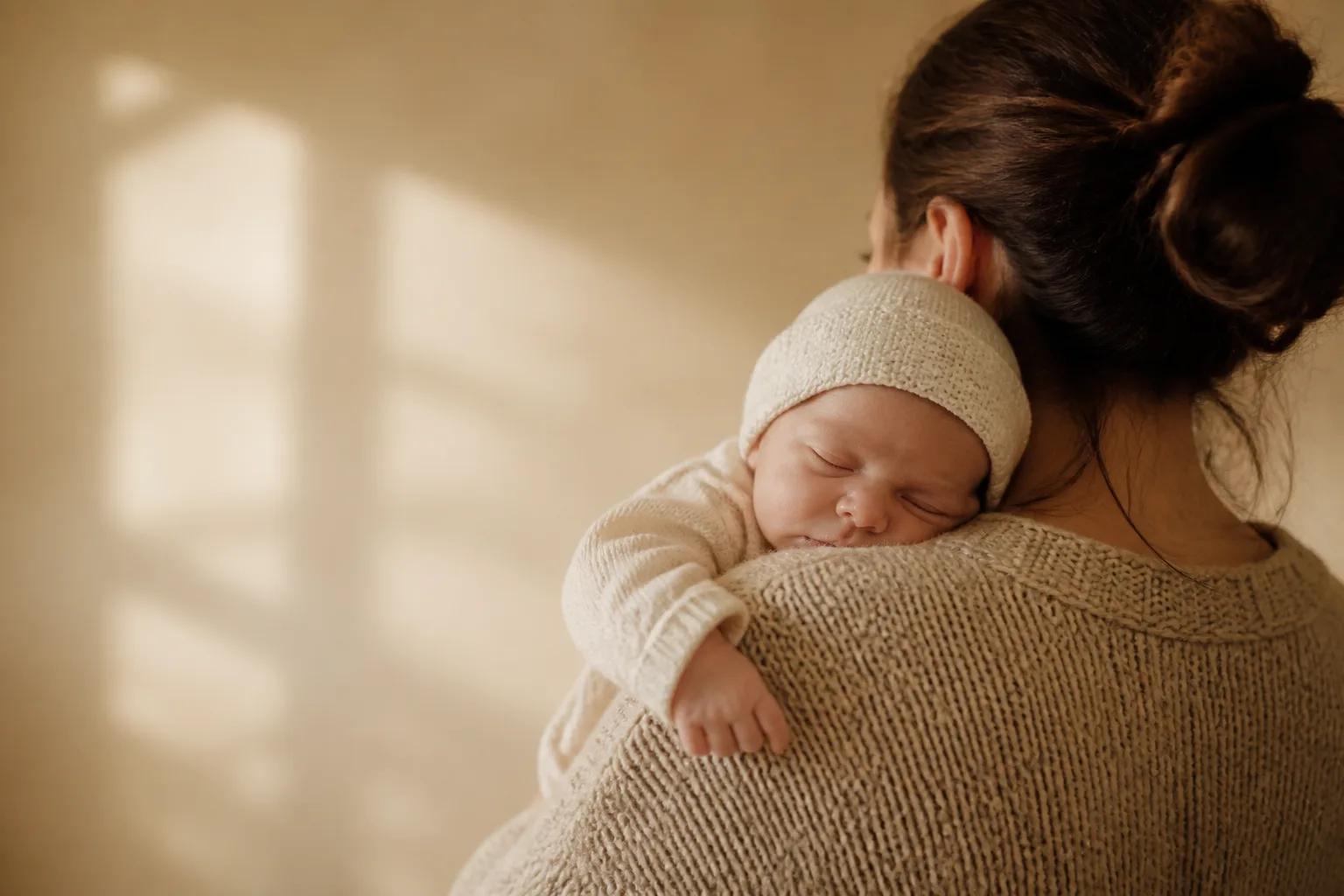 A newborn asleep against a parent's shoulder, seen from behind the parent: the baby in a cream sleepsuit with a fine knit hat, a soft cardigan layered over