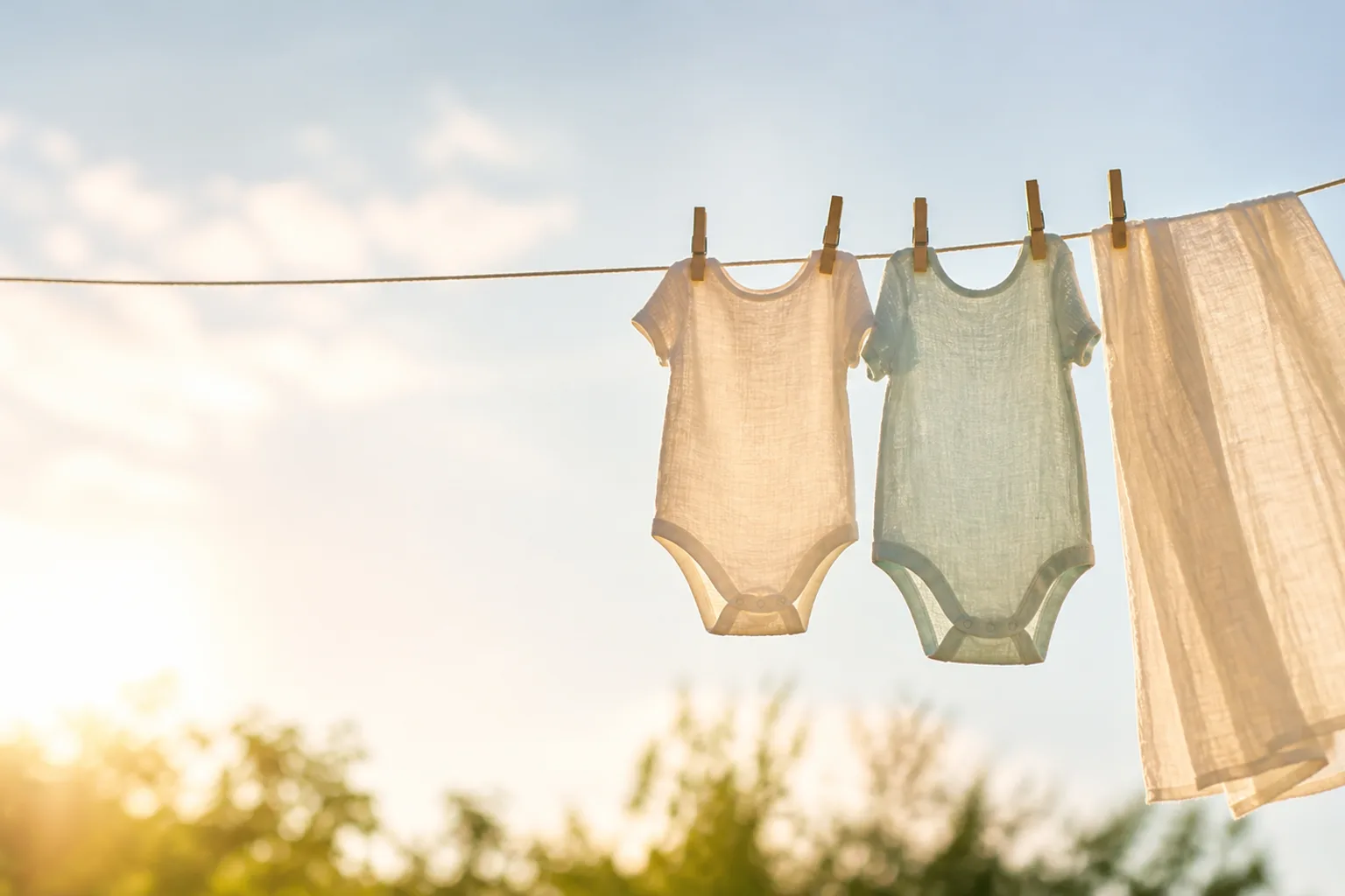Tiny newborn muslin bodysuits and a light muslin swaddle clipped to a clothesline, summer sun glowing through the thin fabric
